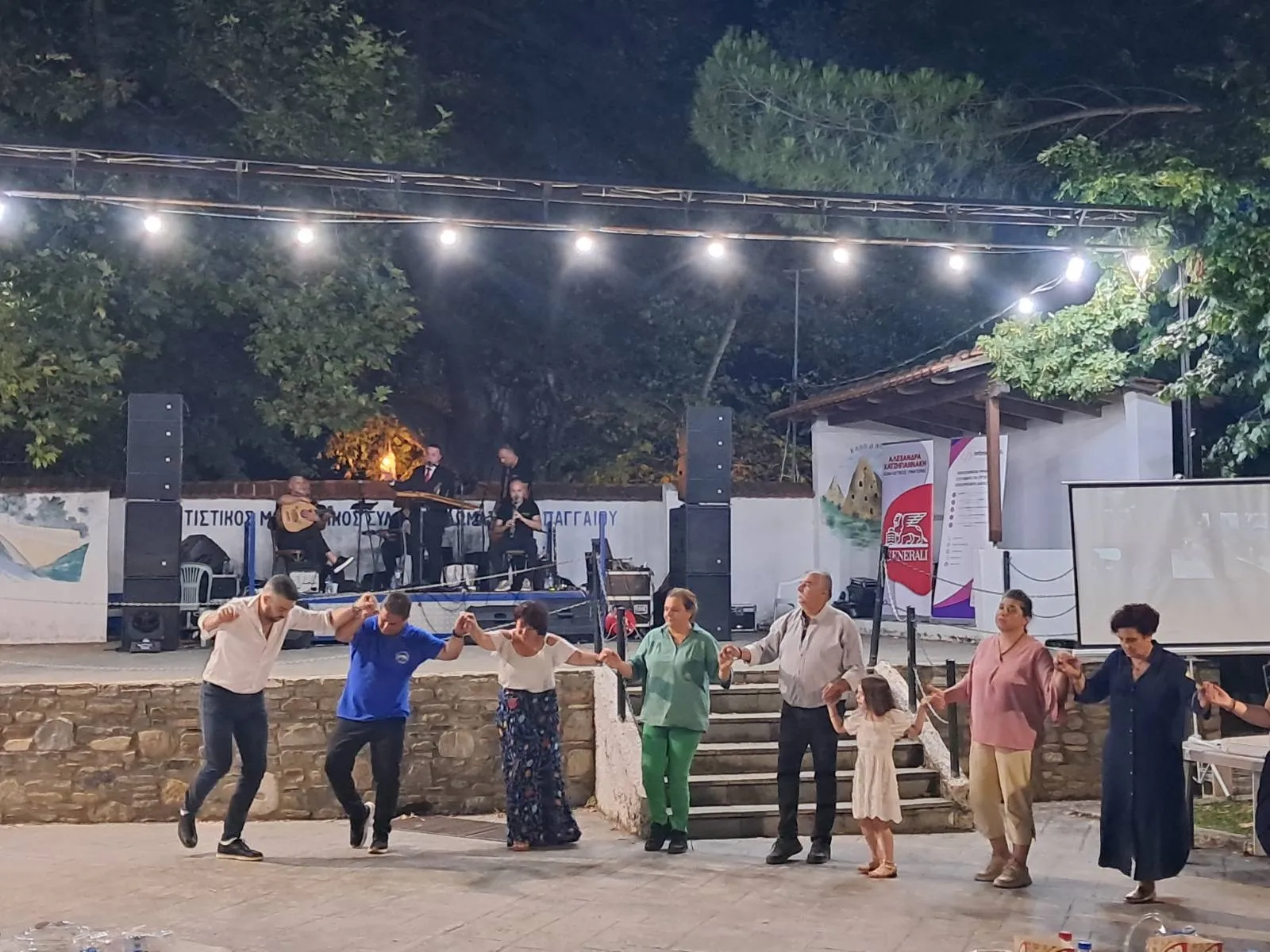 Χωριανοί κάθε ηλικίας χορεύουν πιασμένοι στη σειρά μπροστά από τη σκηνή